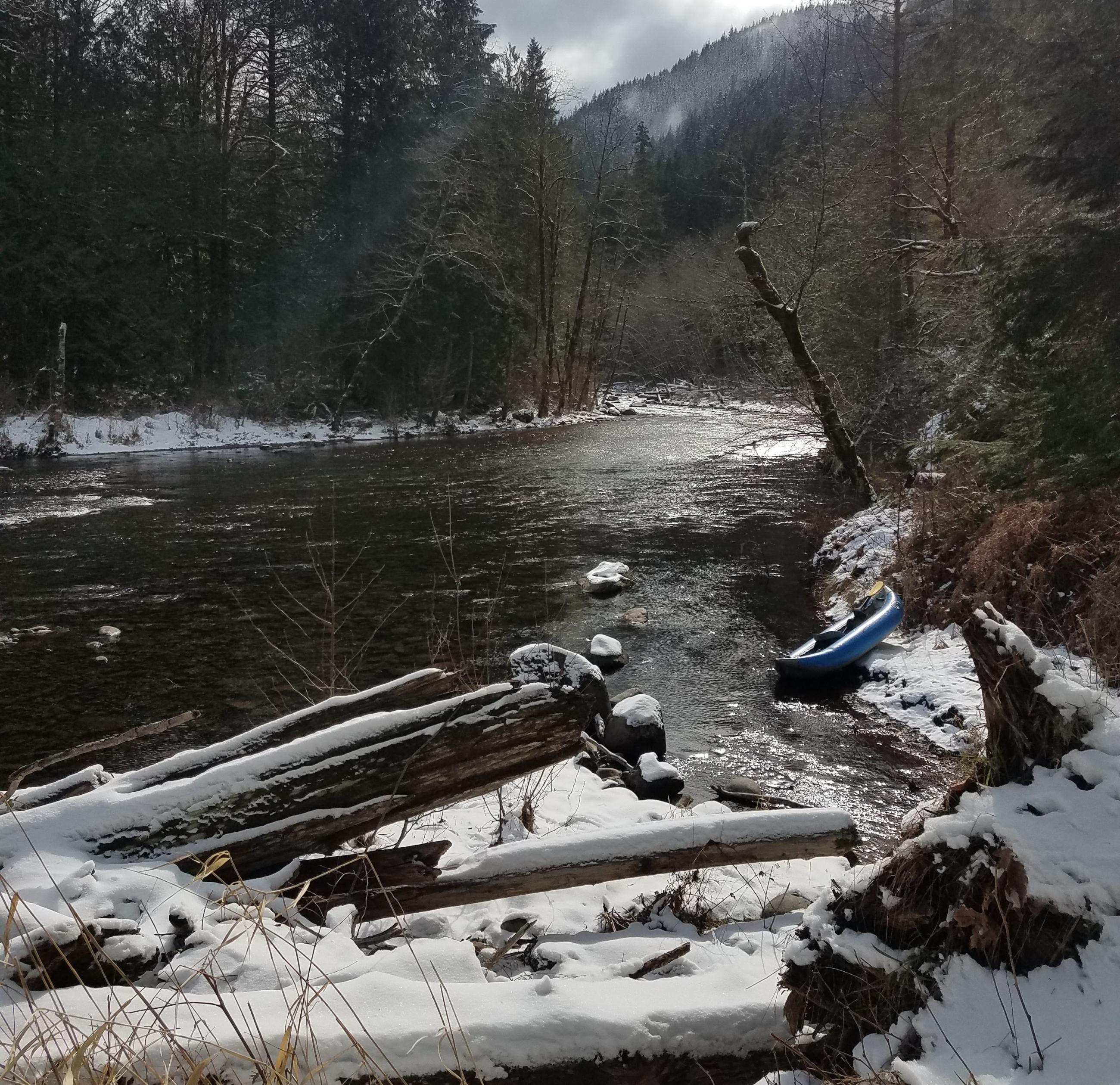 River with bank covered in snow, small blue boat on the right bank with mountains in the background