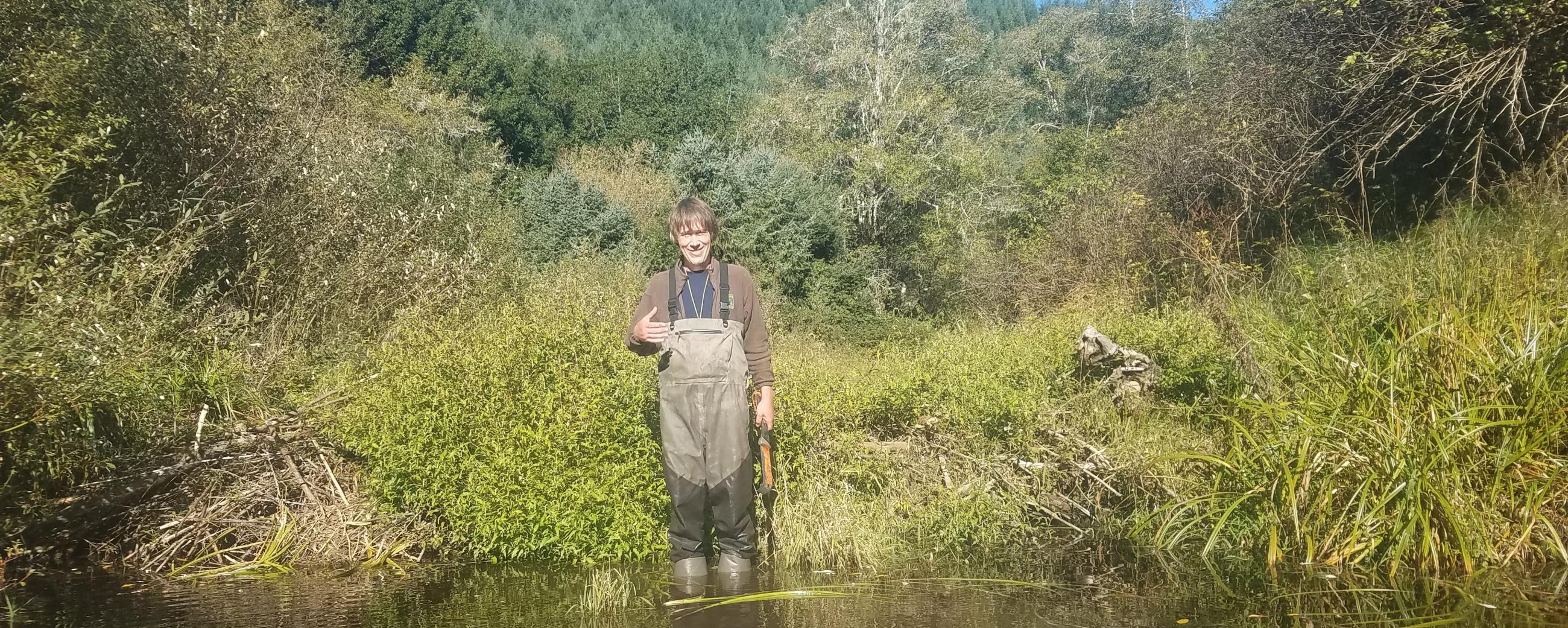 Surveyor stands in waders while holding a machete. He is standing in river water up to his ankles. Behind him is a grassy field and forest scene.