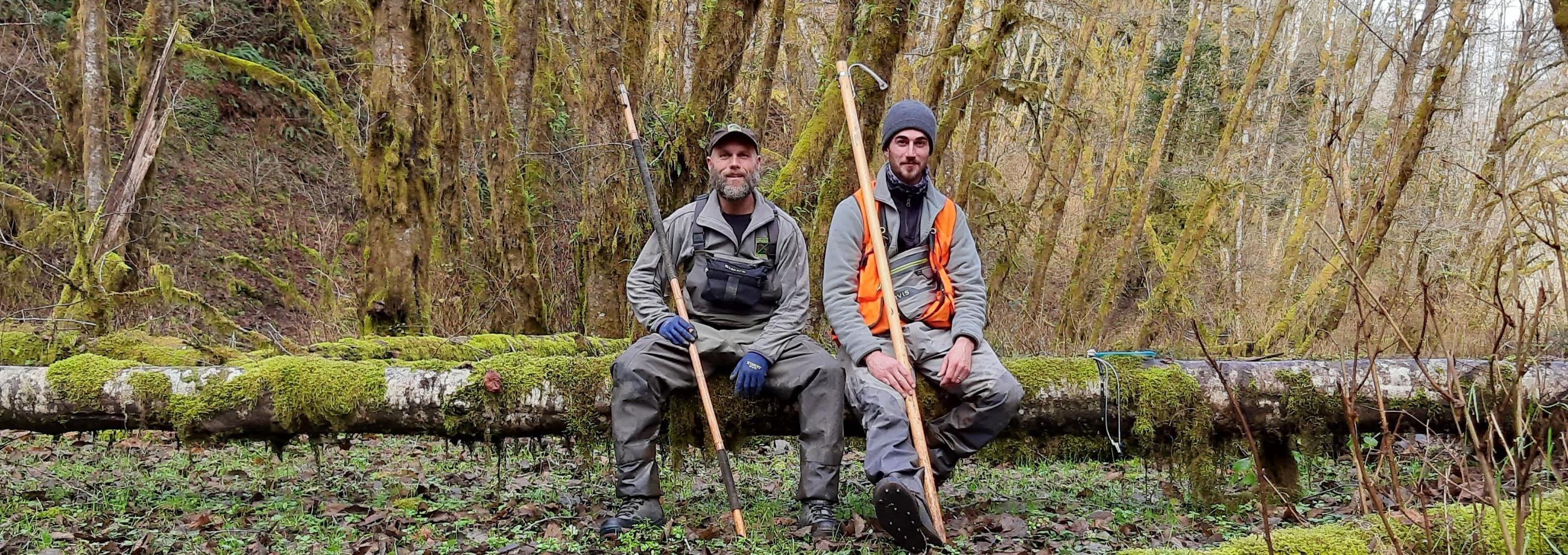 Two male surveyors smile while sitting on a log in the forest.
